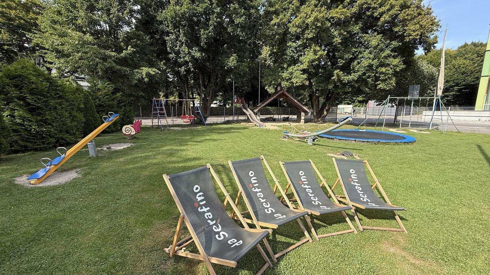 Playground in the greenery with seesaw, swings and trampoline, four deckchairs with the da Serafino logo in front