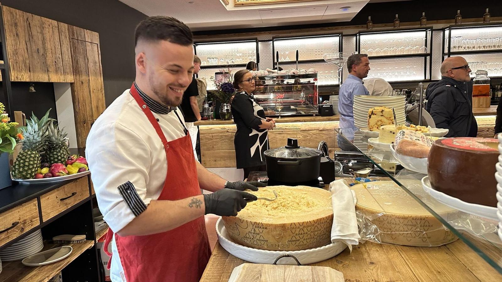 Koch mit roter Schürze schabt lachend Parmesan aus dem großen Käselaib