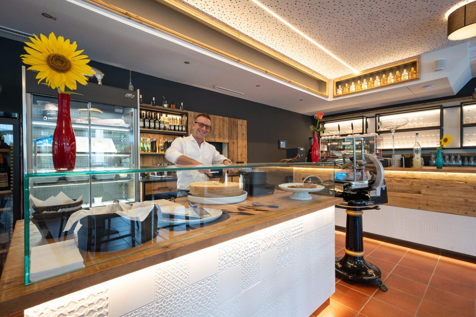 The host smiling behind the counter with a wheel of Parmesan, the illuminated bar in the background