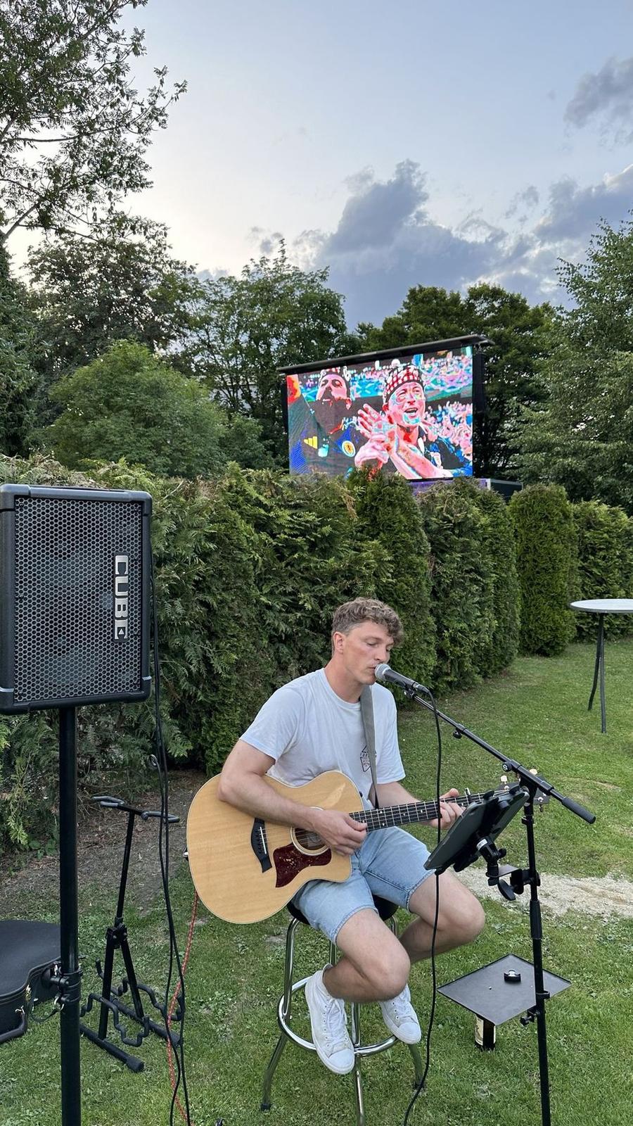 Musiker mit Akustikgitarre singt im Garten, im Hintergrund die Leinwand